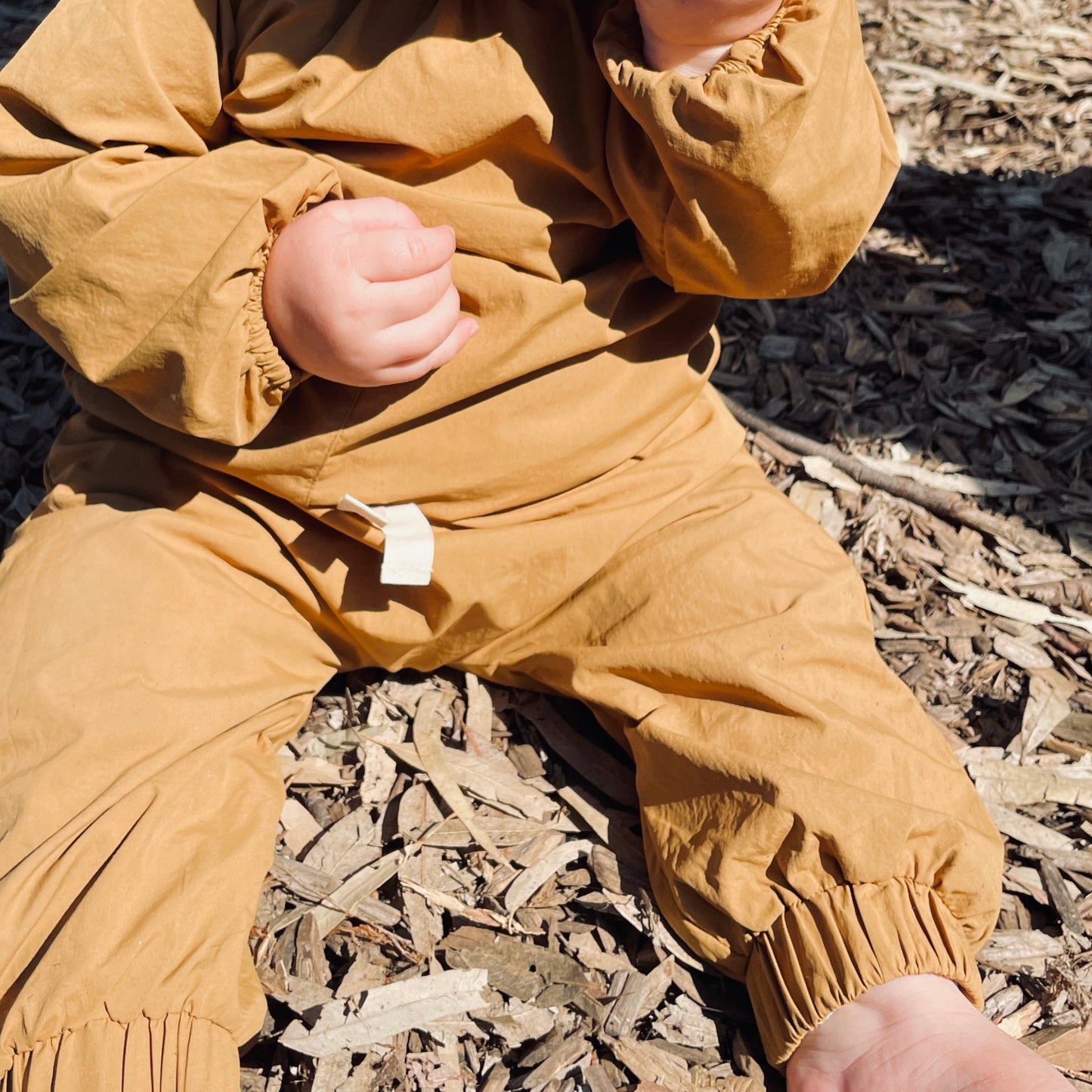 toddler sitting on tan bark eating and exploring at the park in waterproof protective clothes